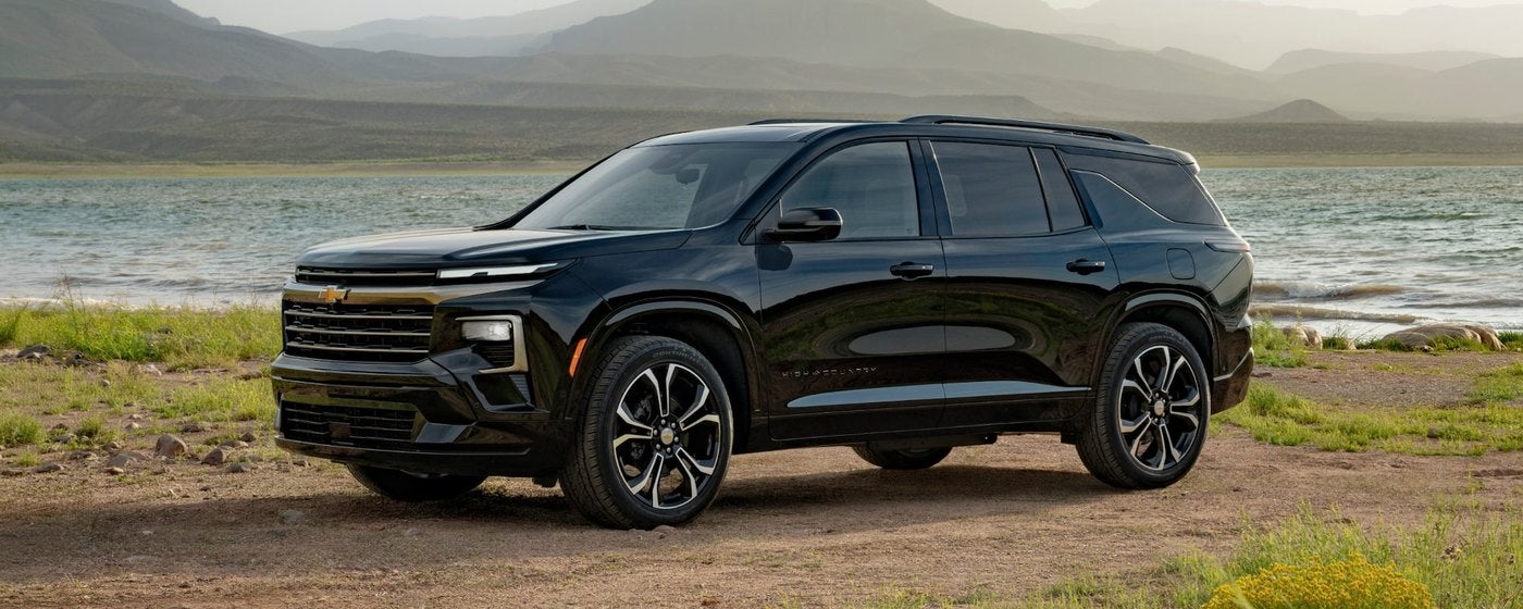 Black Chevrolet Traverse parked on grass beside a lake with hills behind it