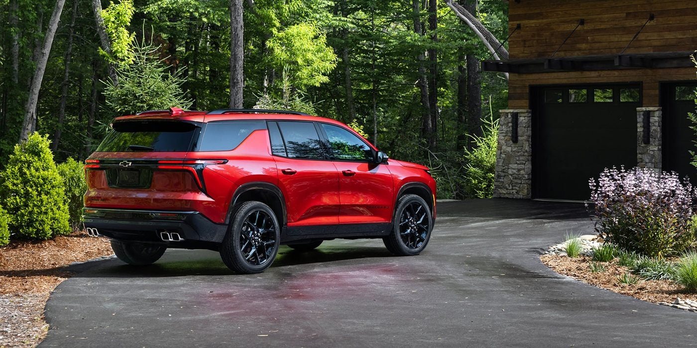 Red Chevrolet Traverse parked on a driveway near a wooded house, seen from the rear