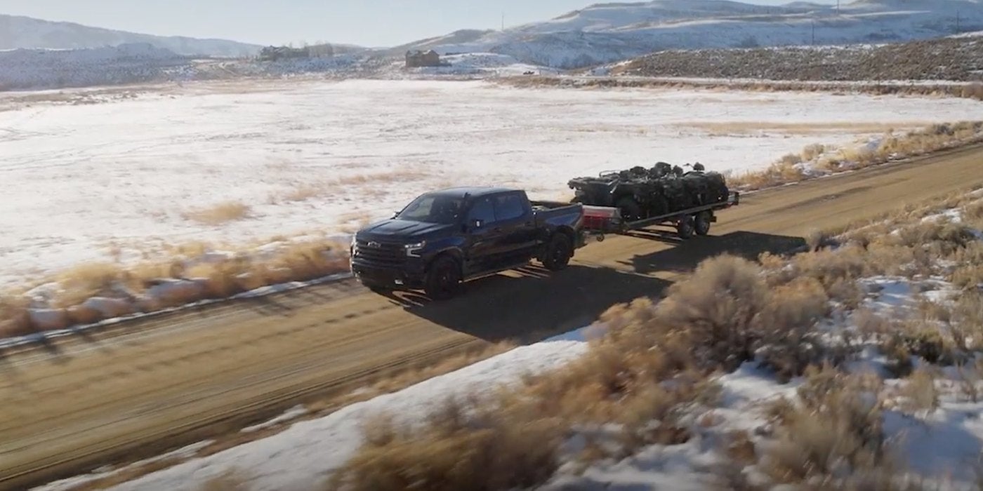 Dark Silverado 1500 pulling a loaded trailer along a dirt road beside a snowy field