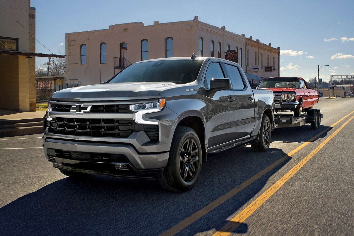 Gray Silverado 1500 towing a classic car on a trailer down a small town main street